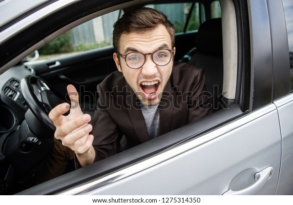 Furious young\
man is looking at the camera while sitting at his car. He is\
screaming at someone. Angry driver\
concept.