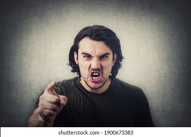 Furious young man pointing forefinger to camera blaming someone as guilty, or scolding isolated on grey wall background. Angry guy screaming and showing with index being annoyed