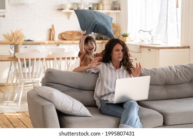 Furious young woman try to concentrate on work as small disobedient kid hit her with pillow. Angry mother freelancer or distance office worker on lockdown at home with hyperactive preschooler child - Shutterstock ID 1995807431