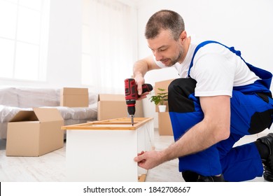 Furniture Assembly. Handyman In Coverall Uniform Assembling And Installing Shelf Using Electric Drill Screwdriver Working Indoors. Carpentry And Carpenter's Service, Repairman Working Fixing Furniture