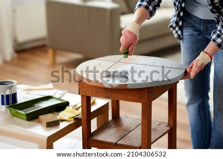 furniture renovation, diy and home improvement concept - close up of woman in gloves with paint roller painting old wooden table in grey color