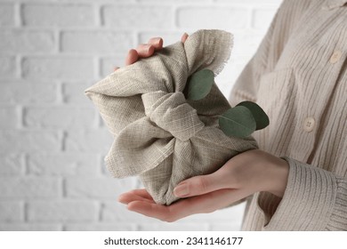 Furoshiki technique. Woman holding gift packed in fabric and decorated with eucalyptus branch against white wall, closeup