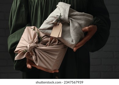 Furoshiki technique. Woman holding gifts wrapped in fabric with thuja branches near dark brick wall, closeup