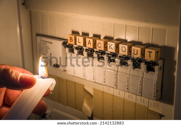 Fuse box with fuses in a distribution box during\
a power outage lit with white candle holding a man with the word\
blackout as text, Germany