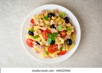 Fusilli Nudelsalat mit Thunfisch, Tomaten, schwarze Oliven und Basilikum auf grauem Hintergrund. Draufsicht.