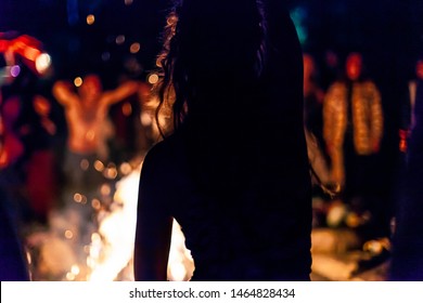 Fusion of cultural & modern music event. A silhouetted girl is seen from behind as she dances by a campfire during a music festival, blurry people and warm glow provides backlight by night
