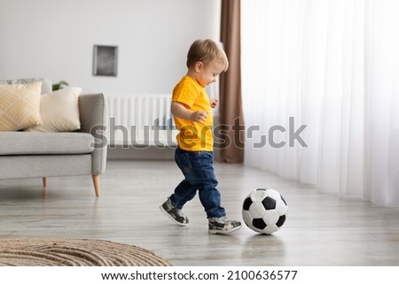 Future champion. Adorable little toddler boy playing football, hitting ball at home, having fun in living room interior, copy space
