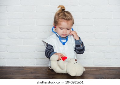 Future profession concept. Baby veterinarian examine toy animal with stethoscope. Game, development, imagination. Childhood, playroom, nursery. Child play doctor with teddy bear on white wall.