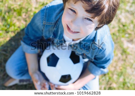 Future professional football player. Selective focus on a face of an adorable preteen kid looking into the camera while sitting in the grass and holding a soccer ball tightly.