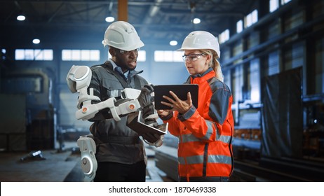 Futuristic Concept Of A Manual Labor Worker In A Bionic Exoskeleton Prototype Working In A Factory. Heavy Industry Engineer Monitors The Powered Suit On An African American Assistant.