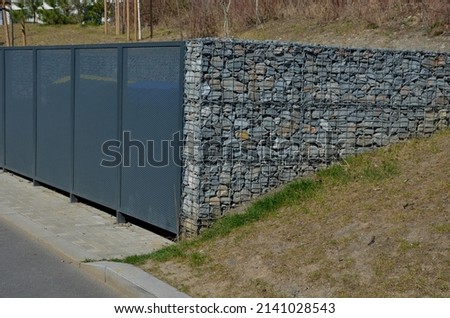 gabion yard for placing containers for sorting household waste. gray door and halving of perforated metal grid, expanded metal. stone wire baskets filled and sunk into grassy slope. reclining place