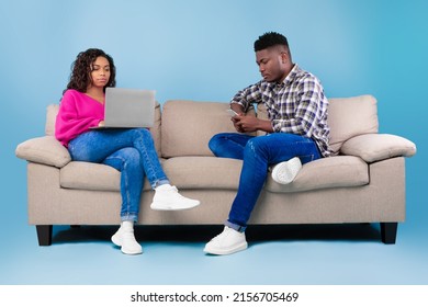 Gadget Addiction. African American Man And Woman Sitting On Couch, Using Mobile Phone And Laptop On Blue Studio Background. Young Black Couple Browsing Internet Instead Of Talking To Each Other