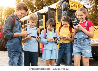 Gadget addiction in children. Diverse preteen kids using smartphones while standing near yellow school bus outdoors, multiethnic boys and girls playing mobile games, scrolling, browsing internet - Powered by Shutterstock