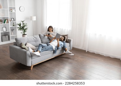 Gadget addiction, modern technologies and millennials lifestyle. Beautiful mixed race spouses resting on sofa at home, using digital tablet and smartphone, high angle view, copy space - Powered by Shutterstock