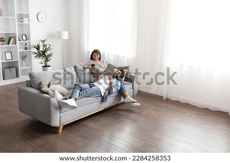 Gadget addiction, modern technologies and millennials lifestyle. Beautiful mixed race spouses resting on sofa at home, using digital tablet and smartphone, high angle view, copy space
