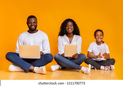 Gadget generation. Happy african american family holding different electronic devices in hands, sitting in a row over yellow studio background