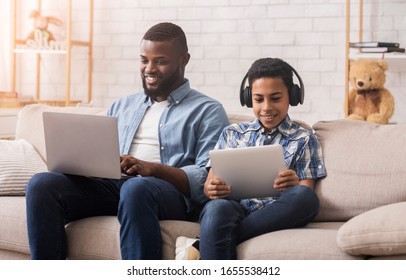 Gadget Generations. Afro father and son spending time with different electronic devices at home, man using laptop and boy holding digital tablet - Powered by Shutterstock