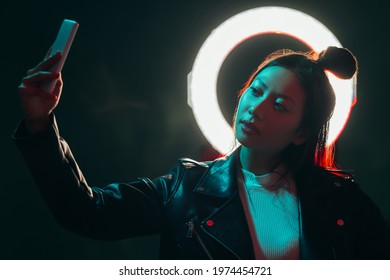 Gadget people. Cyberpunk portrait. Futuristic technology. Blue red neon color light Asian girl in black leather jacket taking selfie on phone with white halo in smoke at dark night. - Powered by Shutterstock