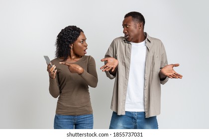 Gadget and social media addiction, cheating in relationships. Black woman checking her boyfriend mobile phone, showing provocative messages and yelling at man, gray studio background