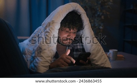 Gadget social media phone addict Indian guy Hispanic man Arabian male sleep disorder insomnia smartphone addiction at night evening home under blanket cover smiling say wow browsing cellphone news app