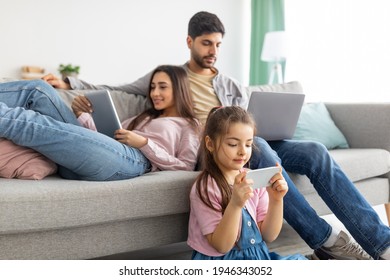 Gadgets Addiction. Eastern Family Using Different Electronic Devices, Resting Together In Living Room, Parents Sitting On Sofa While Their Daughter On Floor. Modern Technologies, Phubbing Concept