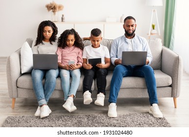 Gadgets Addiction. Portrait Of African American Family Of Four People Holding And Using Different Electronic Devices While Sitting On The Sofa In Living Room At Home, Parents And Their Little Children