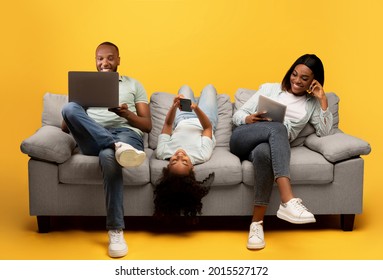 Gadgets addiction. Young black family of three holding and using different electronic devices while sitting on sofa on yellow background. Parents and their daughter with modern gadgets - Shutterstock ID 2015527172