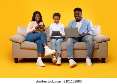 Gadgets Addiction. Young Black Family Of Three People Holding And Using Different Electronic Devices Sitting On Sofa On Yellow Orange Background. Parents And Their Daughter With Modern Technology Pc