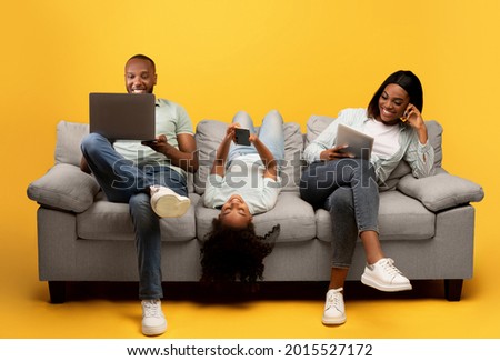 Gadgets addiction. Young black family of three holding and using different electronic devices while sitting on sofa on yellow background. Parents and their daughter with modern gadgets