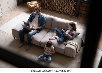 Gadgets addiction. Young family of three holding and using different electronic devices while sitting on sofa in the living room. Father and their daughters with modern gadgets - Powered by Shutterstock