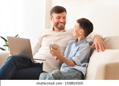 Gadgets. Happy Father And Son Using Phone And Laptop Spending Time Together Sitting On Couch At Home. Selective Focus