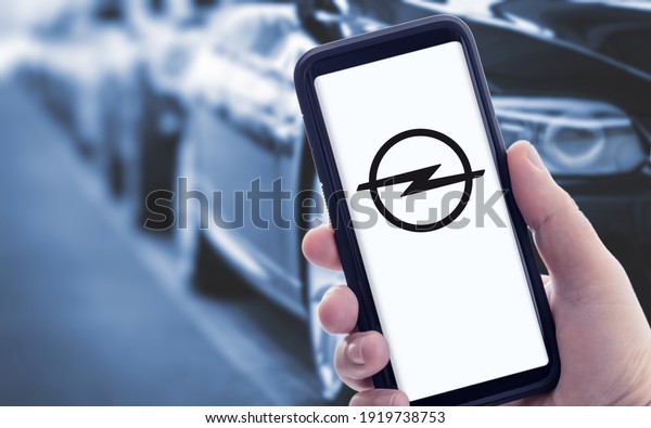 Galicia, Spain; january 09 2021: Hand holding a\
smart phone with new OPEL logo on screen and blurry cars on\
background. Copy space