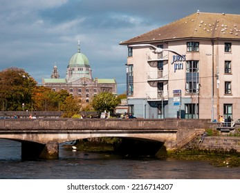 Galway, Ireland - 10.02.2022: Wolfe Tone Bridge And Seagull Sun Bathing On The Roof Of Jurys Inn, Galway Cathedral In The Background. Warm Sunny Day And Blue Cloudy Sky. Town Central Area.