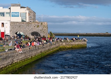 Galway, Ireland - July, 12. 2022: crowds of people sitting on the Spanish arch at Claddagh and enjoying beautiful sunny summer day