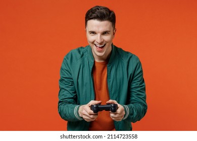 Gambling cheerful fun friendly fascinating happy young brunet man 20s wear red t-shirt green jacket hold in hand play pc game with joystick console isolated on plain orange background studio portrait.