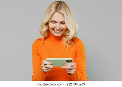 Gambling Elderly Smiling Cool Happy Blonde Woman 50s In Orange Turtleneck Using Play Racing App On Mobile Cell Phone Hold Gadget Smartphone For Pc Video Games Isolated On Plain Grey Background Studio