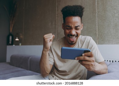Gambling excited young african american man in t-shirt sit on grey soft sofa indoors apartment play racing app mobile cell phone smartphone for pc video games rest stay at home. Tattoo translate fun - Shutterstock ID 2016888128
