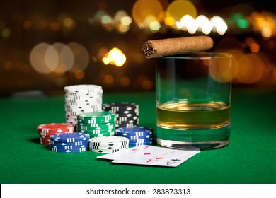 gambling, fortune and entertainment concept - close up of casino chips, whisky glass, playing cards and cigar on green table surface over holidays night lights background