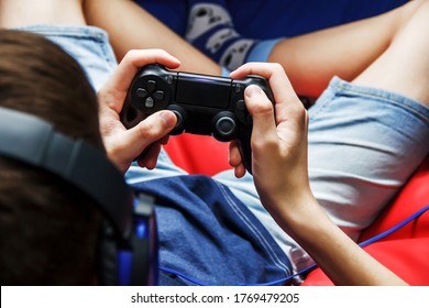 Game Joystick In The Hands Of A Teenager Close-up. Teenage Boy In Big Headphones Sits With A Joystick At Home, Top View. The Guy Is Sitting On A Red Bean Bag Chair. Computer Games. Play Online.