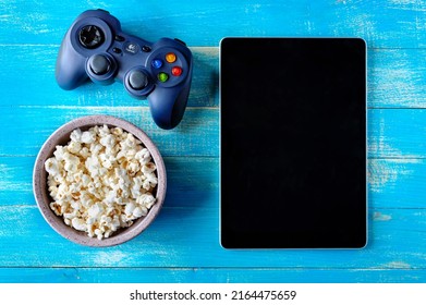 Gamepad LOGITECH, bowl of popcorn and tablet on blue wooden background with copy space. Top view. The concept of leisure, games, pastime.