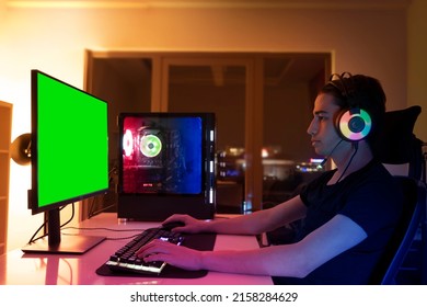 Gamer Boy, Concentrated Gamer Boy In Headset With A Mic, Playing Online Game On His Personal Desktop Computer Or Pc With Green Screen Mockup. Teen Room With Warm RGB Colors, Boy Alone In His Room.