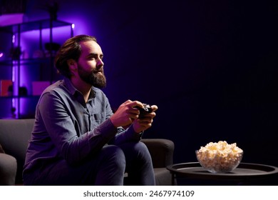 Gamer engaging in online competitive multiplayer tourney using cutting edge gaming console and gamepad while eating popcorn. Man in home theatre playing videogames and consuming snacks - Powered by Shutterstock