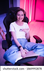 Gamer Girl In A Big Professional Music Headphones In White T-shirt And Blue Jeans Holds Smart Phone In Hands And Plays Game App On Mobile Phone, Smiling And Looking At Camera Indoors.