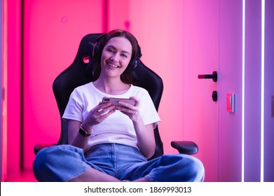 Gamer Girl In A Big Professional Music Headphones In White T-shirt And Blue Jeans Holds Smart Phone In Hands And Plays Game App On Mobile Phone, Smiling And Looking At Camera Indoors.