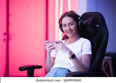 Gamer Girl In A Big Professional Music Headphones In White T-shirt And Blue Jeans Holds Smart Phone In Hands And Plays Game App On Mobile Phone, Smiling And Looking At Camera Indoors.