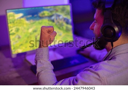 A gamer guy with a fist clenched in a moment of triumph or intense gameplay, engaged with the computer screen