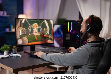 Gamer With Headset Playing Video Games On Computer. Man Wearing Headphones, Using Keyboard And Mousepad To Play Online Video Game On Monitor With Gaming Equipment. Modern Player