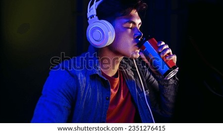Gamer taking a well-deserved break, sipping on a can of energy drink to recharge. In the middle of the night, he savors the refreshing liquid, ready to jump back into his gameplay with renewed energy.