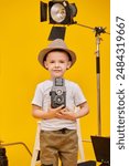 Games in the profession. A cute little boy plays movie director while sitting at a film set on a yellow studio background. Children
