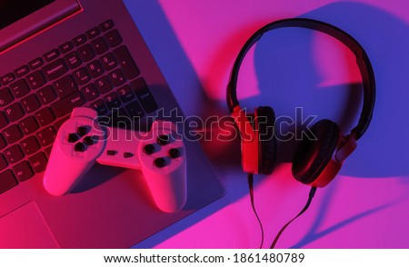 Gaming concept. Gaming equipment. Laptop with gamepad, headphones. Neon red-blue gradient light. Top view